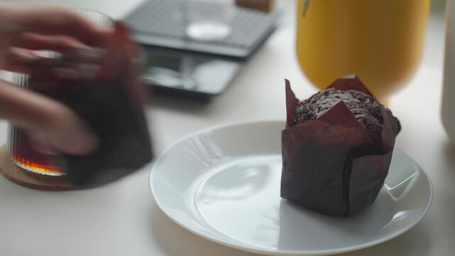 Hand placing chocolate muffin on plate beside glass of coffee. Concept of coffee break and breakfast dessert