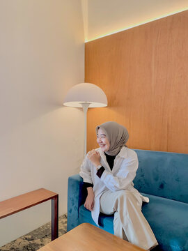 Muslim Woman in White Outerwear and Beige Hijab Sitting on Modern Sofa Indoors. Muslim woman wearing a beige hijab and white outerwear while sitting on a modern sofa indoors, showcasing authentic mode