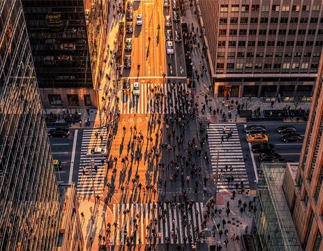 High-angle drone view of business people and commuters crossing a busy city intersection between tall skyscrapers, with dramatic long shadows cast by the golden evening sunset light.
