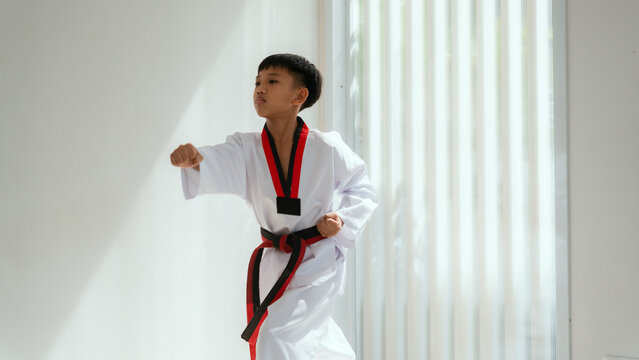 Young boy practices martial arts in a bright room during the day while wearing a taekwondo uniform and holding a black belt