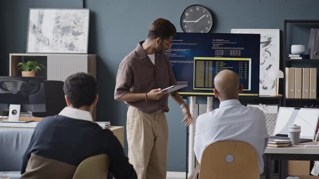 Handheld shot of young Indian male developer presenting code on screen to multiethnic colleagues in IT office. Perfect for software development, coding bootcamp, and tech startup marketing