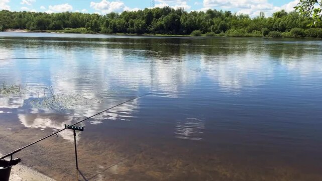 A peaceful scene of recreational fishing on a wide river during a sunny summer day. The calm water reflects white clouds and blue sky, with a lush green forest on the opposite shore
