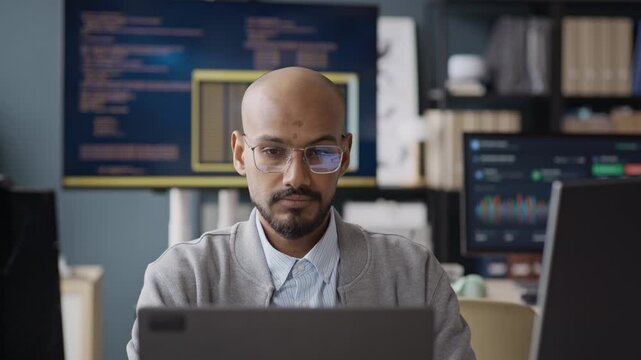 Chest up shot of focused Indian young male software engineer wearing glasses coding on laptop in IT office. Footage is perfect for software development, data science, tech recruitment campaigns