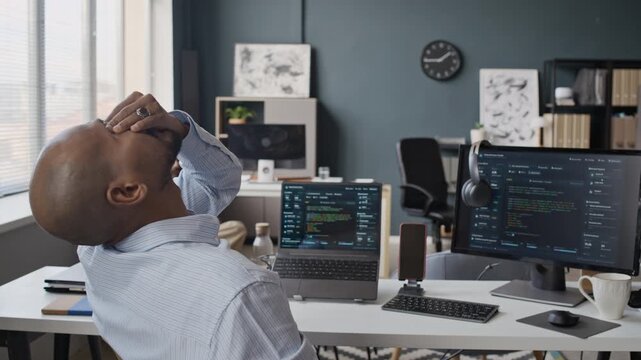 Handheld zoom out shot of young Indian male employee taking off glasses and rubbing eyes in exhaustion while coding on computer in IT office. For campaigns on mental health and workplace stress