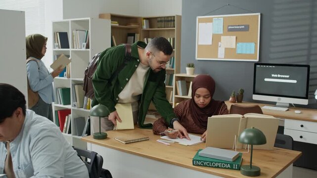 Diverse young adult students studying in library, Caucasian male student explaining assignment to Muslim female student wearing hijab. Clip suitable for promoting inclusive higher education