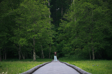 Fototapeta premium Forest boardwalk woman meadow tree green nature summer, lone woman standing on wooden path through lush woodland and grassy meadow with calm expression