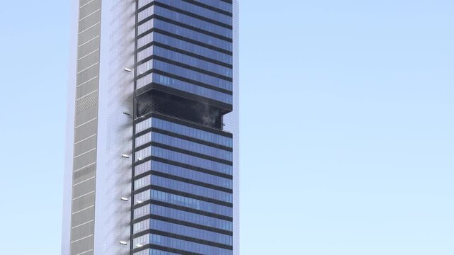 Dangerous fire burning on an upper floor of a modern office skyscraper against a clear blue sky