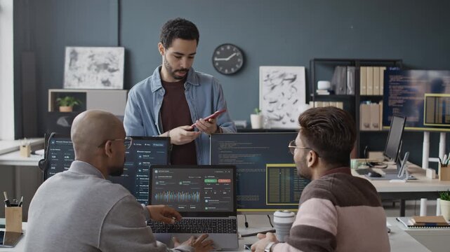 Handheld shot of diverse young male developers discussing mobile app design while looking at smartphone and laptop screens in office. Perfect for tech startup marketing and software engineering ads