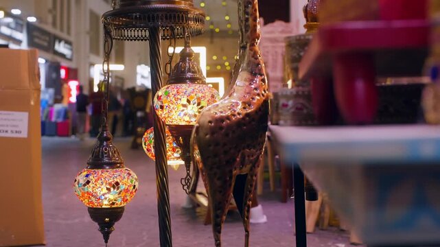 Decorative Turkish style lanterns with colorful glass mosaics glow warmly while suspended in crowded bazaar or marketplace, creating festive atmosphere with blurred shoppers, lights in background.