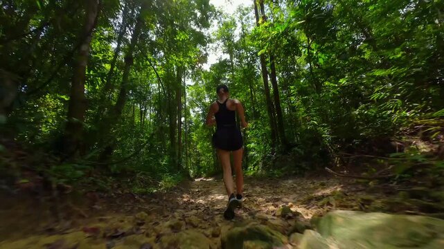 Active woman jogging on a forest trail surrounded by dense tropical rainforest. Low angle FPV drone tracking shot showcasing outdoor fitness, adventure and healthy lifestyle in in Southeast Asia,