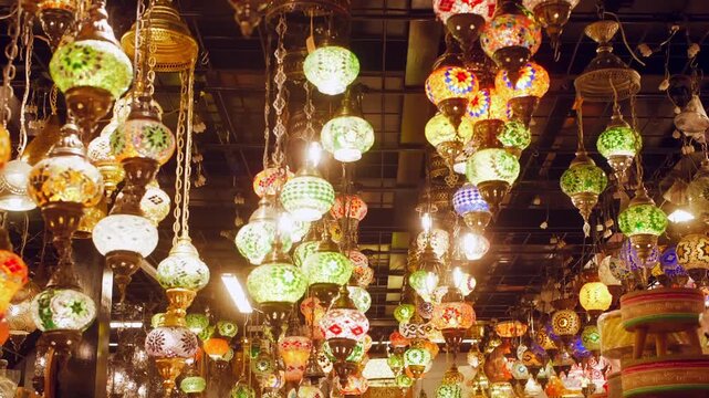 Rows of decorative glass lanterns with intricate brass fittings hang from ceiling chains in vibrant colors including green, orange, white, creating warm ambient lighting Middle Eastern bazaar setting.