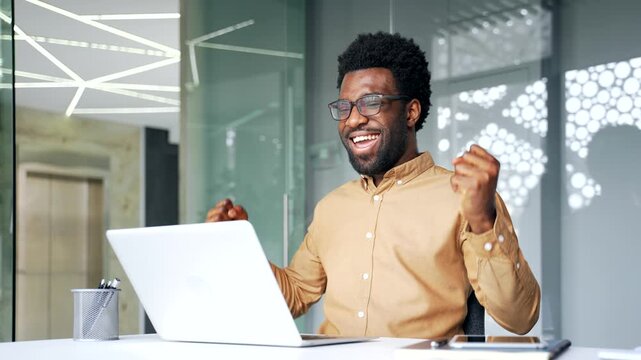 Shocked and surprised African American businessman smiling while celebrating financial success on laptop in office. Excited black man manager reads positive good message, happy with achievement.