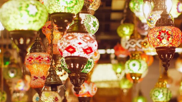 Multiple decorative lanterns with intricate colored glass panels hang from ceiling in warm golden lighting, creating festive atmospheric display in traditional Middle Eastern bazaar setting.