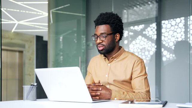 Confident African American businessman sitting at workplace desk and talking on video call using laptop. Successful black man manager speaking at online conference during professional business meeting