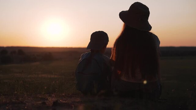 Mother and child sit watching sunset in countryside field. Woman with boy enjoys golden hour outdoors. Family sits at sunset together. Mother and child bond in nature. Boy with mother at dusk.