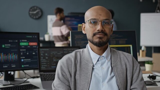 Chest up slowmo portrait of Indian young male developer in glasses posing for camera in modern IT office. Ideal for tech recruitment, software development services, and corporate IT promos