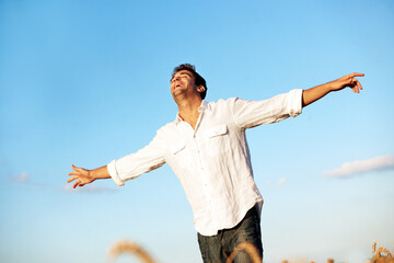 Obraz premium Freedom, happy and man in countryside with blue sky, gratitude and open arms for peace on weekend. Field, smile and person with joy, wellness and relax for holiday, break and adventure outdoor