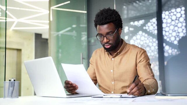Focused African American businessman holding document and writing notes at desk. Serious black man financier checks business information and fills out corporate paper form inside office room.