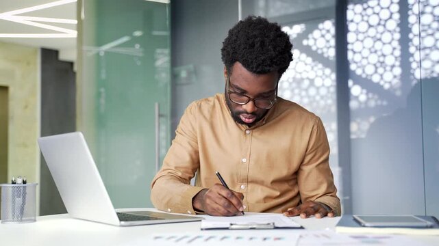 Focused African American businessman signs paper contract at desk. Serious black man financier sitting at workplace inside corporate office puts signature on important financial document.