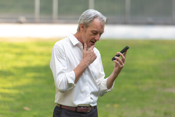 Fototapeta premium Senior adult professional business man holding smartphone, talking business and standing outdoor in a park among office buildings.