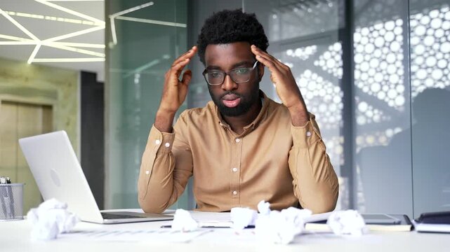 Bored African American businessman sits at office desk with crumpled papers and feels deeply tired. Overworked financier inside corporate workspace experiences severe depression and exhaustion.