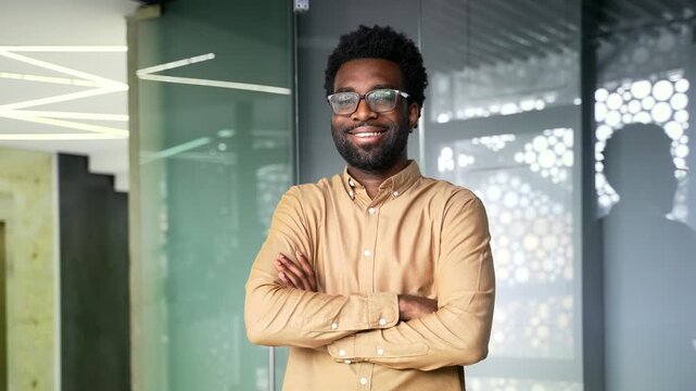 Portrait of happy African American businessman standing at modern office workplace. Successful black man manager wearing glasses looks at camera and poses with crossed arms inside corporate workspace.