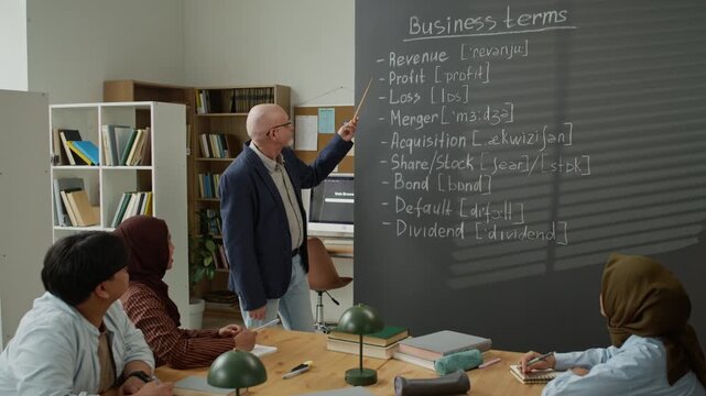 Senior Caucasian teacher explaining business terms on blackboard to diverse young adult migrant students. Clip is ideal for promoting adult education, language schools, and integration programs