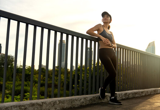 Wide shot of Asian sport girl lean on rail and look to her right side with smiling with warm light of evening.