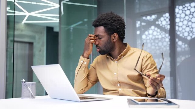 Tired African American businessman has severe eye strain at work. Exhausted black man worker sitting at corporate office desk takes off glasses and rubs bridge of nose because of headache indoors.