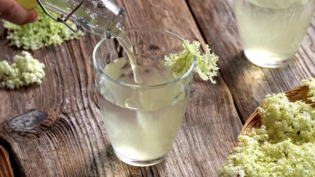 Pouring homemade elderberry flower syrup into a glass cup with water to prepare healthy natural lemonade