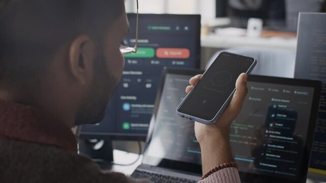 Close up slowmo shot of young Indian man using smartphone while working at computer desk in IT office. Clip is perfect for cybersecurity and mobile app development commercials