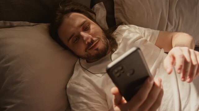 Caucasian young man lying in bed, scrolling on smartphone, writing messages and browsing social media. Clip is ideal for mobile app marketing, digital addiction, or insomnia concepts