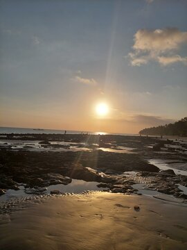 Golden Sunset Over a Rocky Beach:
The bright sun sets slowly behind the distant dark hills in the background. Meanwhile, scattered dark rocks rest heavily across the wet, reflective sand in the immed