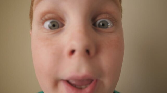 Little red-haired boy looking into camera with wow expression indoor. Portrait of ginger child with freckles showing surprise and amazement. Close up shocked male face of small kid inside. Slow motion