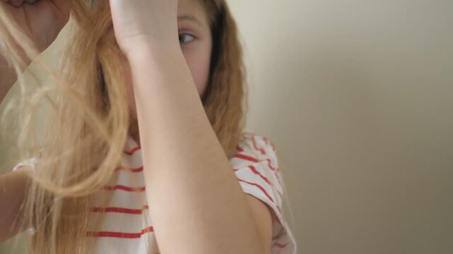 Close up of beautiful small girl weaving small pigtail out of her long blonde hair indoor. Little cute female child doing herself a hairstyle at home. Slow motion