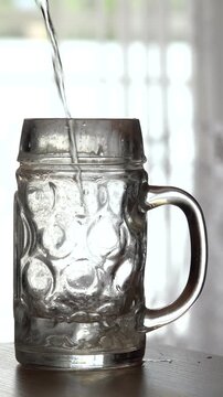 Ein leeres Glas auf einem Tisch vor einem hellen Fenster wird mit Wasser gef&uuml;llt