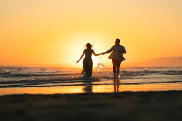Fototapeta premium Couple runs along the beach at sunset while splashing in the water near the waves
