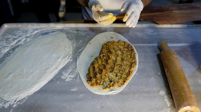 Close-up of gloved hands spreading minced meat mixture over dough during lahmacun preparation. Traditional Turkish food making, kitchen work, savory topping process and handmade meal production.