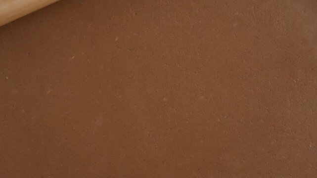 Baker rolls out raw gingerbread cookie dough texture with wooden rolling pin. Pastry chef prepares sweet chocolate dessert recipe background for holiday biscuit baking production process.