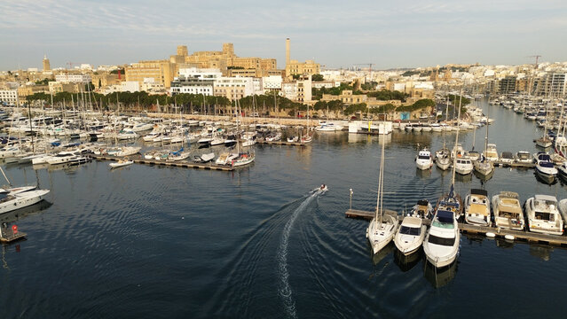 Aerial view of Msida Marina filled with numerous yachts and boats, with a small motorboat creating a wake in the calm water, set against the urban skyline of Malta.