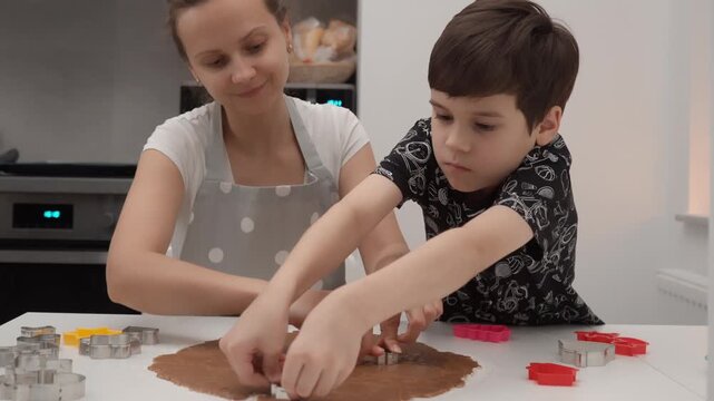 Mother and son cut shapes from gingerbread dough with metal cutters at kitchen table. Family prepares sweet homemade cookies for holiday baking recipe education lifestyle leisure context.