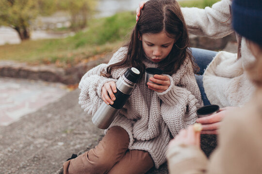 Young girl blowing on hot drink from thermos during outdoor picnic with her family. Family connection, comfort and care in nature, childhood, travel, seasonal lifestyle,.
