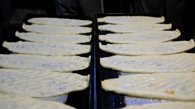 Prepared pides lined up on black baking trays in a bakery kitchen, ready for the oven. Food production, tray arrangement, baking preparation and professional kitchen workflow concept.
