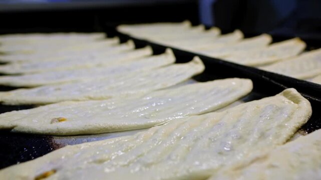Prepared pides lined up on black baking trays in a bakery kitchen, ready for the oven. Food production, tray arrangement, baking preparation and professional kitchen workflow concept.