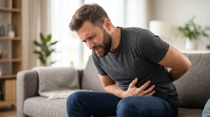 Fototapeta premium A man sits on a couch holding his stomach with an expression of discomfort. He looks concerned while sitting in a well-lit living room with plants and a cozy atmosphere