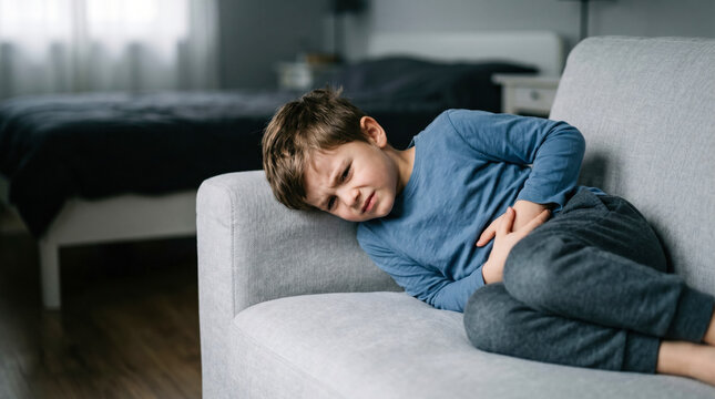 A young boy sits on a couch with a pained expression. He is holding his stomach and looks uncomfortable. The room has soft light coming in