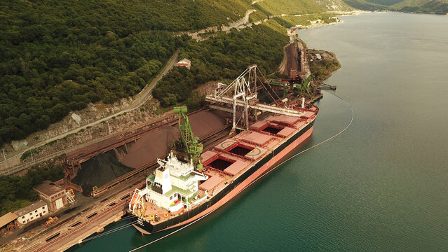 Aerial view of a large bulk carrier ship docked at a port facility, with industrial cranes loading cargo near a forested coastline in Bakarac, Primorje-Gorski Kotar, Croatia.
