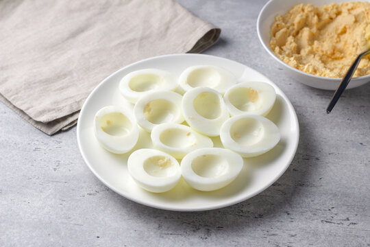 Hard boiled egg halves with yolks removed on a white plate, prepared for stuffed eggs recipe on a gray textured surface with soft natural light