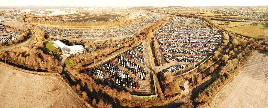 Aerial view of a large new car storage facility filled with rows of parked vehicles, surrounded by rural landscape and industrial infrastructure in Gloucestershire, United Kingdom.