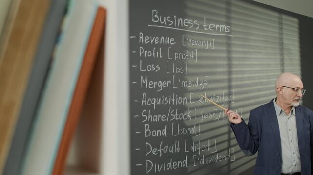 Senior Caucasian male teacher pointing at blackboard with business terms and phonetic transcriptions, explaining pronunciation. Footage is perfect for language school ads and adult education